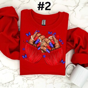 May include: Red long-sleeve shirt with a graphic of two hands with blue and red nail polish, rings, and butterflies. The hands are clasped together in a heart shape. The shirt has the number "#2" printed on the top.