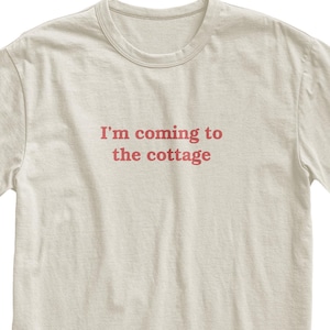 Könnte beinhalten: Cremefarbenes T-Shirt mit dem roten Aufdruck "I'm coming to the cottage". Das Shirt hat kurze Ärmel und einen Rundhalsausschnitt. Der Text ist mittig auf der Brust platziert.