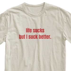 Op de afbeelding: Een beige t-shirt met de rode tekst "life sucks but I suck better." Het shirt met korte mouwen heeft een ronde hals. De tekst staat in het midden op de voorkant van het kledingstuk.