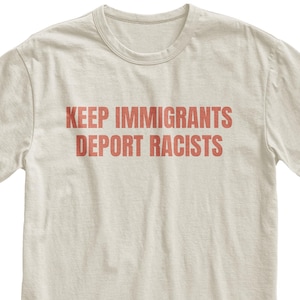 May include: A cream-colored t-shirt with the text "KEEP IMMIGRANTS DEPORT RACISTS" printed in red. The shirt has a crew neck and short sleeves. The text is centered on the front of the shirt.