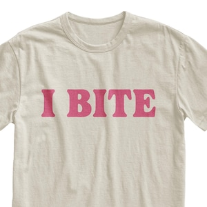 May include: Cream-colored t-shirt with the text "I BITE" in a bold, pink font. The short-sleeved shirt has a crew neck and a simple, casual design. The text is centered on the front of the shirt.