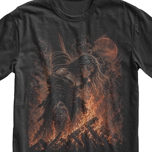 May include: A black short-sleeve t-shirt featuring a graphic print of a figure tied to a wooden post amidst flames. The illustration depicts a dark, fiery scene with orange and red embers, smoke, and a large, glowing red moon in the background.