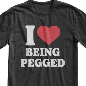 May include: A black short-sleeved crew neck t-shirt featuring a graphic print on the chest. The design includes a white letter I, a red heart symbol, and the white text BEING PEGGED arranged in two lines below the heart and letter.