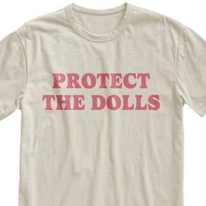 Puede incluir: Una camiseta beige con las palabras "PROTECT THE DOLLS" impresas en rosa.