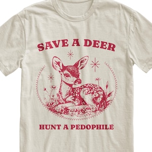 Könnte beinhalten: Cremefarbenes T-Shirt mit roter Grafik eines Rehs, umgeben von Blumen, und dem Text "SAVE A DEER" darüber und "HUNT A PEDOPHILE" darunter. Das Reh liegt.
