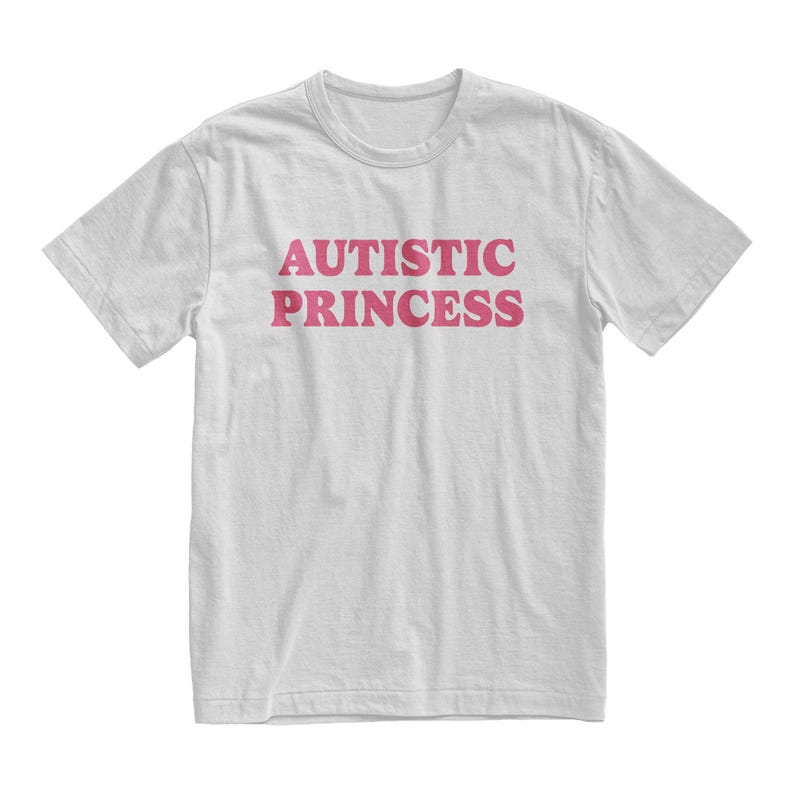 Puede incluir: Camiseta blanca con el texto "AUTISTIC PRINCESS" en rosa, en letras may&uacute;sculas y en negrita. La camiseta tiene mangas cortas y cuello redondo. El texto est&aacute; centrado en la parte delantera.