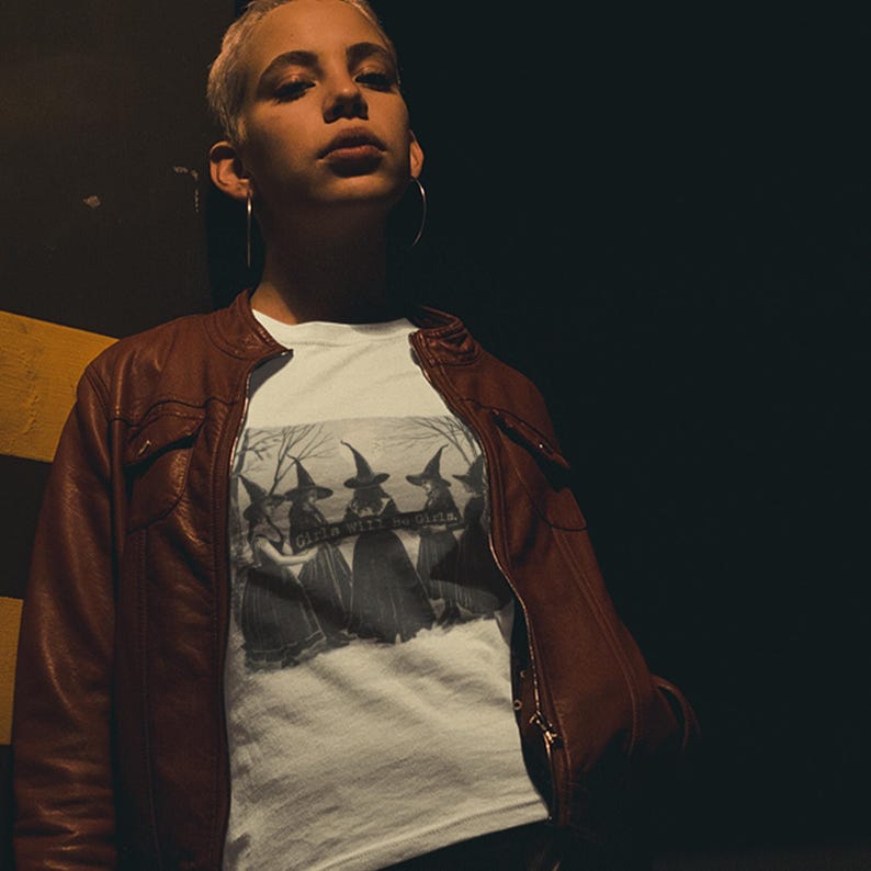 K&ouml;nnte beinhalten: Eine Person tr&auml;gt eine braune Lederjacke &uuml;ber einem wei&szlig;en T-Shirt mit einer schwarz-wei&szlig;en Grafik von f&uuml;nf Hexen mit dem Text "Girls will be girls" darauf.