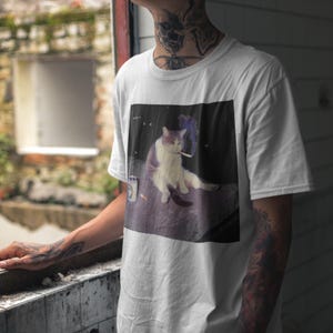 Op de afbeelding: Wit t-shirt met een afbeelding van een kat die een sigaret rookt. De kat is wit en paars, met een sigaret in zijn mond en rook die opstijgt. Een blikje en een sigarettenpeuk zijn ook zichtbaar.