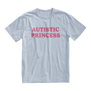 Puede incluir: Camiseta azul claro con la frase "AUTISTIC PRINCESS" en rosa estampada en el frente. La camiseta tiene mangas cortas y cuello redondo.