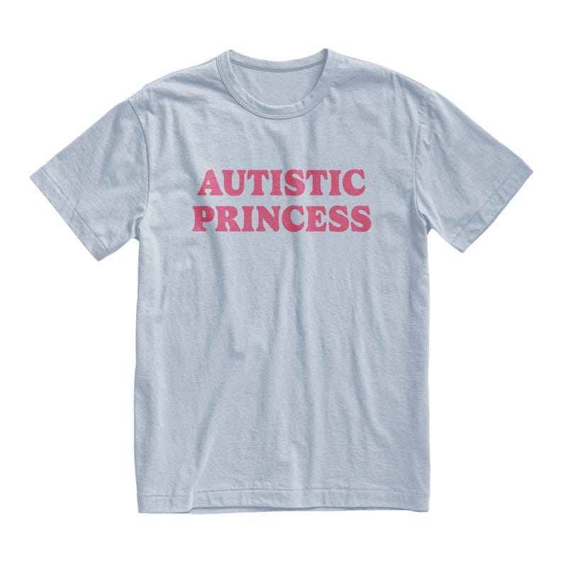 Puede incluir: Camiseta azul claro con la frase "AUTISTIC PRINCESS" en rosa estampada en el frente. La camiseta tiene mangas cortas y cuello redondo.