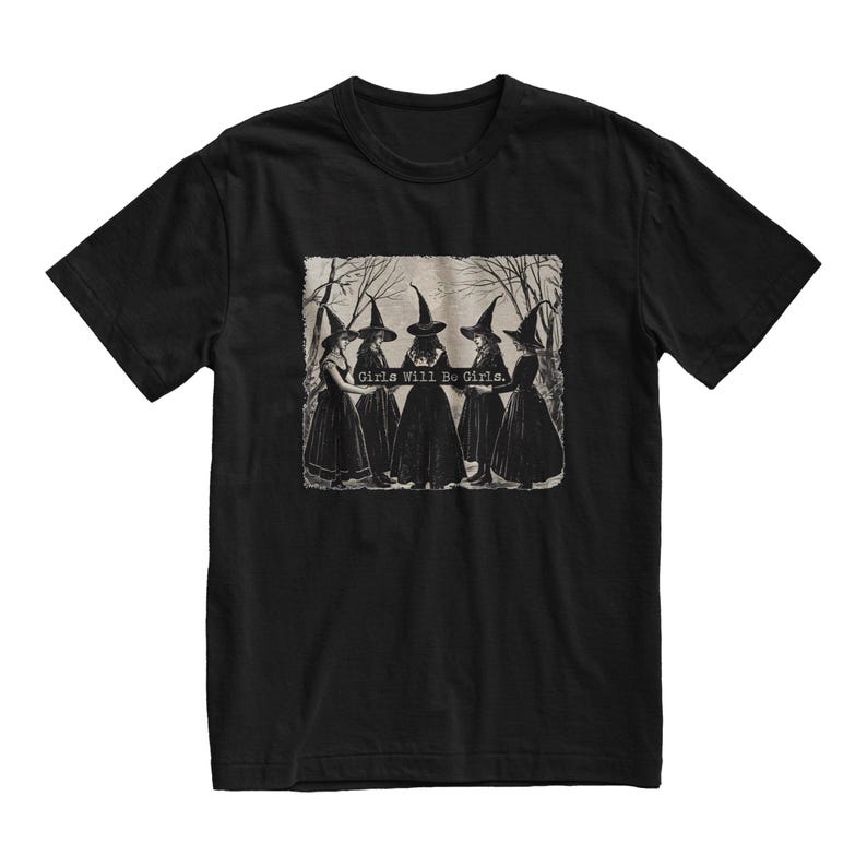 K&ouml;nnte beinhalten: Schwarzes T-Shirt mit einem grafischen Druck von f&uuml;nf Hexen, die schwarze spitze H&uuml;te und Kleider tragen. Der Text "Girls Will Be Girls." ist auf dem Grafikdruck.
