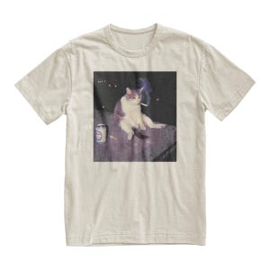 Op de afbeelding: Beige t-shirt met een afbeelding van een kat die een sigaret rookt. De kat is wit en grijs, zittend op een oppervlak met een blikje en een sigaret. De afbeelding heeft een vintage, licht vervaagde uitstraling.