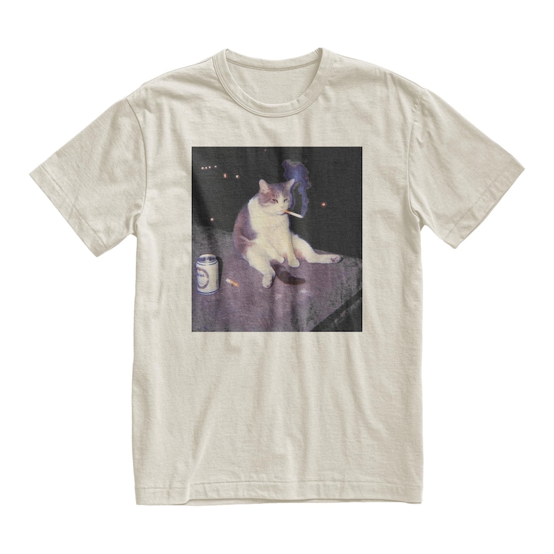 Op de afbeelding: Beige t-shirt met een afbeelding van een kat die een sigaret rookt. De kat is wit en grijs, zittend op een oppervlak met een blikje en een sigaret. De afbeelding heeft een vintage, licht vervaagde uitstraling.