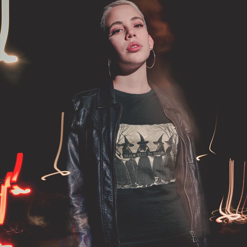K&ouml;nnte beinhalten: Eine Person tr&auml;gt eine schwarze Lederjacke &uuml;ber einem dunkelgr&uuml;nen T-Shirt mit einer Vintage-Illustration von drei Hexen und dem Text "Girls will be girls".