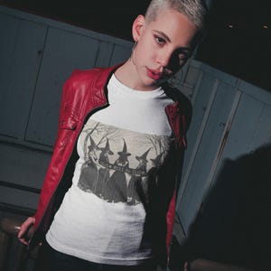 K&ouml;nnte beinhalten: Ein wei&szlig;es T-Shirt mit einer Schwarzwei&szlig;illustration von vier Hexen, die spitze H&uuml;te tragen. Der Text "Girls Will Be Girls." ist auf das Shirt gedruckt.