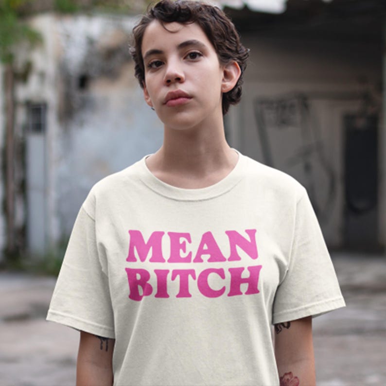 May include: Cream-colored t-shirt with the text "MEAN BITCH" in bold, bubble-style pink letters. The shirt has short sleeves and a crew neck. The person in the image is wearing the shirt.