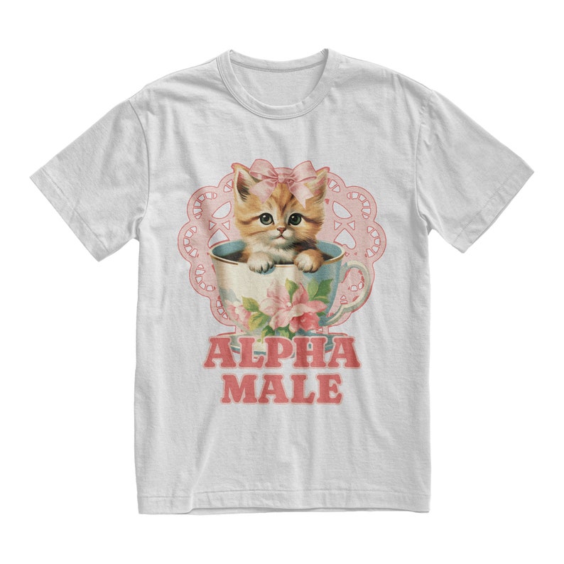 Pu&ograve; includere: Maglietta bianca con un'illustrazione vintage di un gattino in una tazza da t&egrave; con fiori rosa e un pizzo rosa. Il testo "ALPHA MALE" &egrave; stampato in rosa sotto l'immagine.