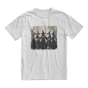 K&ouml;nnte beinhalten: Wei&szlig;es T-Shirt mit einer Schwarzwei&szlig;illustration von f&uuml;nf Hexen, die schwarze spitze H&uuml;te und Kleider tragen. Der Text "Girls Will Be Girls." ist auf das T-Shirt gedruckt.