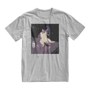 Op de afbeelding: Lichtgrijs t-shirt met een afbeelding van een kat die een sigaret rookt. De kat is wit en grijs, zittend met een blikje en sigaretten in de buurt. De afbeelding heeft een donkere achtergrond met een paar lichten.