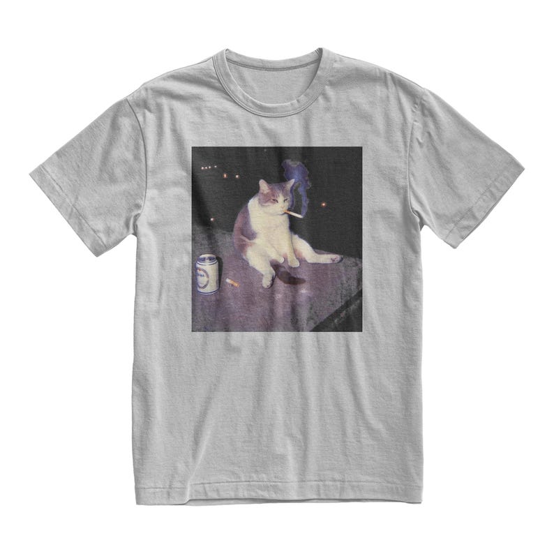 Op de afbeelding: Lichtgrijs t-shirt met een afbeelding van een kat die een sigaret rookt. De kat is wit en grijs, zittend met een blikje en sigaretten in de buurt. De afbeelding heeft een donkere achtergrond met een paar lichten.