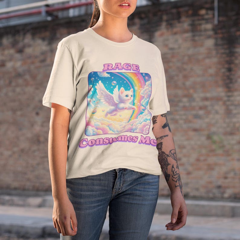 May include: A white t-shirt with a graphic of a white cat with wings flying in a rainbow sky. The text "Rage Consumes Me" is printed in pink below the graphic.