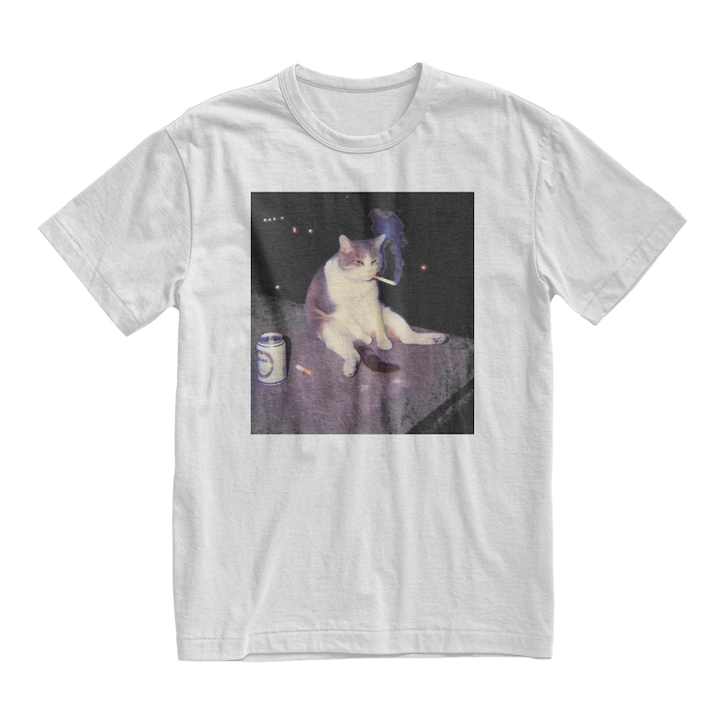 Op de afbeelding: Wit t-shirt met een meme-achtige afbeelding van een kat die een sigaret rookt. De kat wordt afgebeeld met een blikje en een sigaret. De afbeelding heeft een vintage, licht vervaagde esthetiek. Het shirt heeft een klassieke ronde hals.