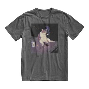 Op de afbeelding: Donkergrijs t-shirt met een meme van een kat die een sigaret rookt. De kat is wit en grijs, zittend op een oppervlak met een blikje en een sigaret in de buurt. De afbeelding heeft een donkere achtergrond met een paar lichten.