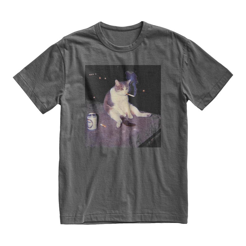 Op de afbeelding: Donkergrijs t-shirt met een meme van een kat die een sigaret rookt. De kat is wit en grijs, zittend op een oppervlak met een blikje en een sigaret in de buurt. De afbeelding heeft een donkere achtergrond met een paar lichten.