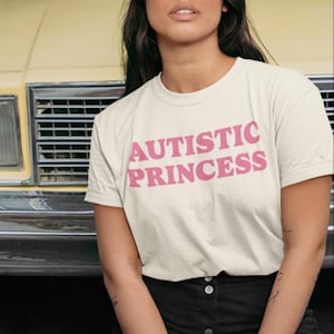 Puede incluir: Camiseta color crema con las palabras "AUTISTIC PRINCESS" en rosa y letras gruesas. La camiseta es de cuello redondo y parece estar hecha de una tela suave. La persona lleva una falda negra.