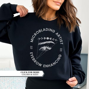 May include: Black sweatshirt with a white circular graphic that reads "MICROBLADING ARTIST EYEBROW ENHANCING" around an eye and eyebrow design. The text "ESTD 23" is also included. The sweatshirt has a crew neck.