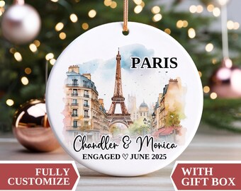 May include: A white ceramic Christmas ornament featuring a watercolor illustration of Paris, including the Eiffel Tower and buildings. The ornament has the word "PARIS" at the top, with the names "Chandler & Monica" and the date "JUNE 2025" below the image.