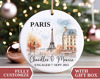 May include: White ceramic Christmas ornament featuring a watercolor illustration of Paris with the Eiffel Tower and a building. Text includes "PARIS", "Chandler & Monica", and "ENGAGED SEPT 2025". Includes a gold string for hanging.