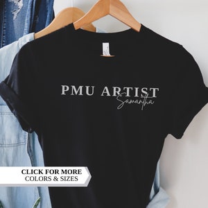 May include: Black t-shirt with white text that reads "PMU ARTIST Samantha" and a white banner that reads "CLICK FOR MORE COLORS & SIZES". The t-shirt is hanging on a wooden hanger. The text "MYRA GRACE CO." is visible in the bottom right corner.