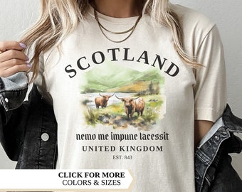 May include: A natural-colored t-shirt with the word "SCOTLAND" in black, above a watercolor image of two brown Highland cows. Below the image is the text "nemo me impune lacessit" and "UNITED KINGDOM EST. 843". The shirt also has the text "CLICK FOR MORE COLORS & SIZES".