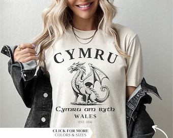 May include: A cream-colored t-shirt featuring a black dragon design with the text "CYMRU" above and "Cymru am Byth WALES EST. 1056" below. The shirt has a crew neck and short sleeves. The words "CLICK FOR MORE COLORS & SIZES" are also visible.