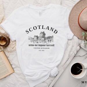 May include: White t-shirt with a black graphic of a Scottish castle and the word "SCOTLAND" above it. The text "nemo me impune lacessit" and "UNITED KINGDOM EST. 843" are below the graphic. A beige hat and a cup of coffee are also in the image.