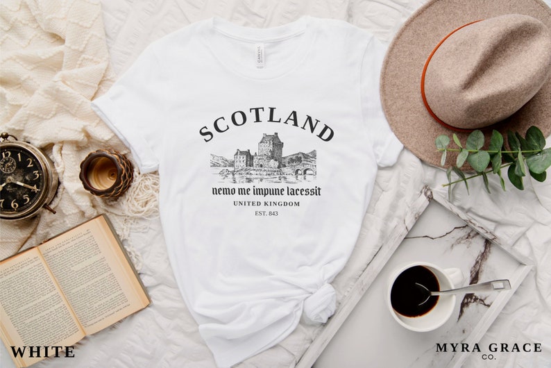 May include: White t-shirt with a black graphic of a Scottish castle and the word "SCOTLAND" above it. The text "nemo me impune lacessit" and "UNITED KINGDOM EST. 843" are below the graphic. A beige hat and a cup of coffee are also in the image.
