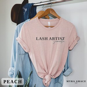 Pu&ograve; includere: Una maglietta color pesca con la scritta "LASH ARTIST Samantha" stampata in nero. La maglietta &egrave; appesa a un gancio di legno con una camicia di jeans blu e un cappello marrone sullo sfondo. La parola "PEACH" &egrave; in una scatola bianca in basso.