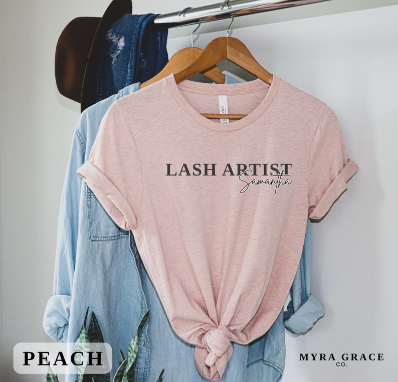 Pu&ograve; includere: Una maglietta color pesca con la scritta "LASH ARTIST Samantha" stampata in nero. La maglietta &egrave; appesa a un gancio di legno con una camicia di jeans blu e un cappello marrone sullo sfondo. La parola "PEACH" &egrave; in una scatola bianca in basso.