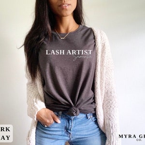 Pu&ograve; includere: T-shirt grigio scuro con la scritta "LASH ARTIST Samantha" in corsivo bianco. La maglietta &egrave; abbinata a jeans blu e un cardigan color crema. Sono visibili anche le parole "DARK GRAY" e "MYRA GRACE CO.".