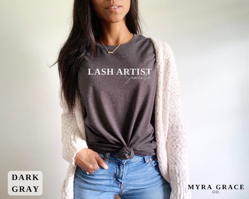Pu&ograve; includere: T-shirt grigio scuro con la scritta "LASH ARTIST Samantha" in corsivo bianco. La maglietta &egrave; abbinata a jeans blu e un cardigan color crema. Sono visibili anche le parole "DARK GRAY" e "MYRA GRACE CO.".