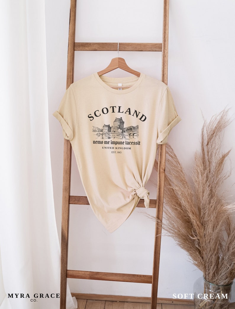 May include: A soft cream-colored t-shirt with the word "SCOTLAND" printed across the top in black, along with a graphic of a castle and the text "nemo me impune lacessit UNITED KINGDOM EST. 843". The shirt is hanging on a wooden ladder.