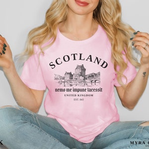 May include: Pink t-shirt with a black graphic of a Scottish castle and the word "SCOTLAND" above it. Below the graphic is the text "nemo me impune lacessit" and "UNITED KINGDOM EST. 843". The shirt is a light pink color.