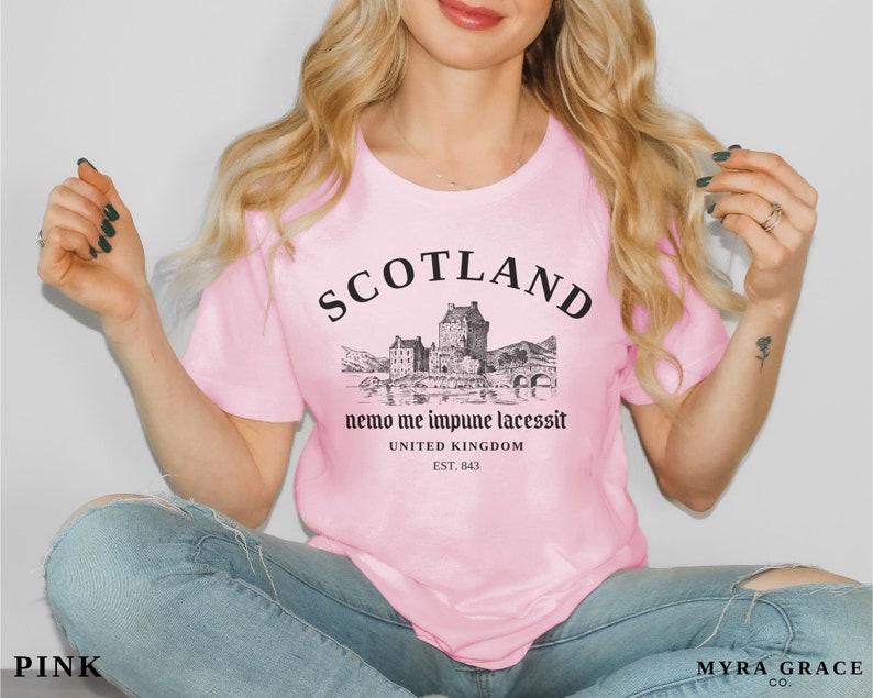 May include: Pink t-shirt with a black graphic of a Scottish castle and the word "SCOTLAND" above it. Below the graphic is the text "nemo me impune lacessit" and "UNITED KINGDOM EST. 843". The shirt is a light pink color.