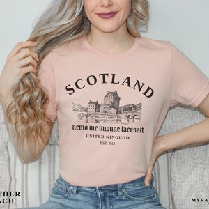 May include: Heather peach-colored t-shirt with a black graphic of a Scottish castle and the word "SCOTLAND" above it. The text "nemo me impune lacessit" and "UNITED KINGDOM EST. 843" are also printed on the shirt.