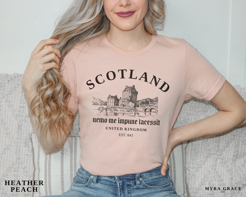 May include: Heather peach-colored t-shirt with a black graphic of a Scottish castle and the word "SCOTLAND" above it. The text "nemo me impune lacessit" and "UNITED KINGDOM EST. 843" are also printed on the shirt.