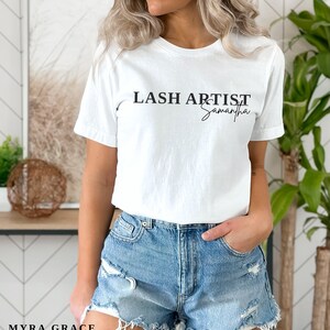 Pu&ograve; includere: T-shirt bianco con la scritta nera "LASH ARTIST Samantha". La maglietta &egrave; abbinata a pantaloncini di jeans. Il logo Myra Grace Co. &egrave; visibile in basso a sinistra nell'immagine. La modella indossa la maglietta.