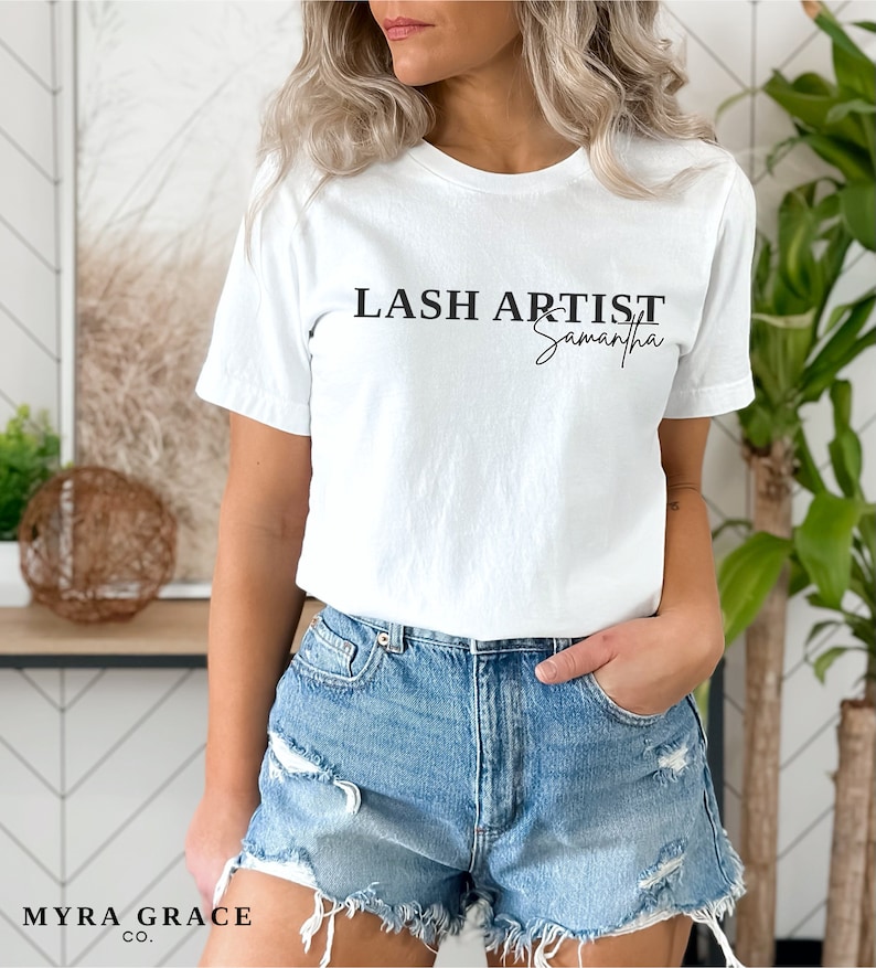 Pu&ograve; includere: T-shirt bianco con la scritta nera "LASH ARTIST Samantha". La maglietta &egrave; abbinata a pantaloncini di jeans. Il logo Myra Grace Co. &egrave; visibile in basso a sinistra nell'immagine. La modella indossa la maglietta.