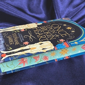 May include: A hardcover book titled "Once Upon a Broken Heart" by Stephanie Garber. The book cover features a blue, green, and gold design with a heart-shaped keyhole and a red and white heart.