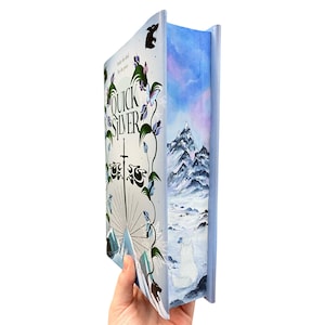 May include: A hardback book titled "Quicksilver" with a fantasy cover. The cover displays a sword, floral designs, and the title in a stylized font. The book's edge features a painted winter landscape with mountains and a white wolf.